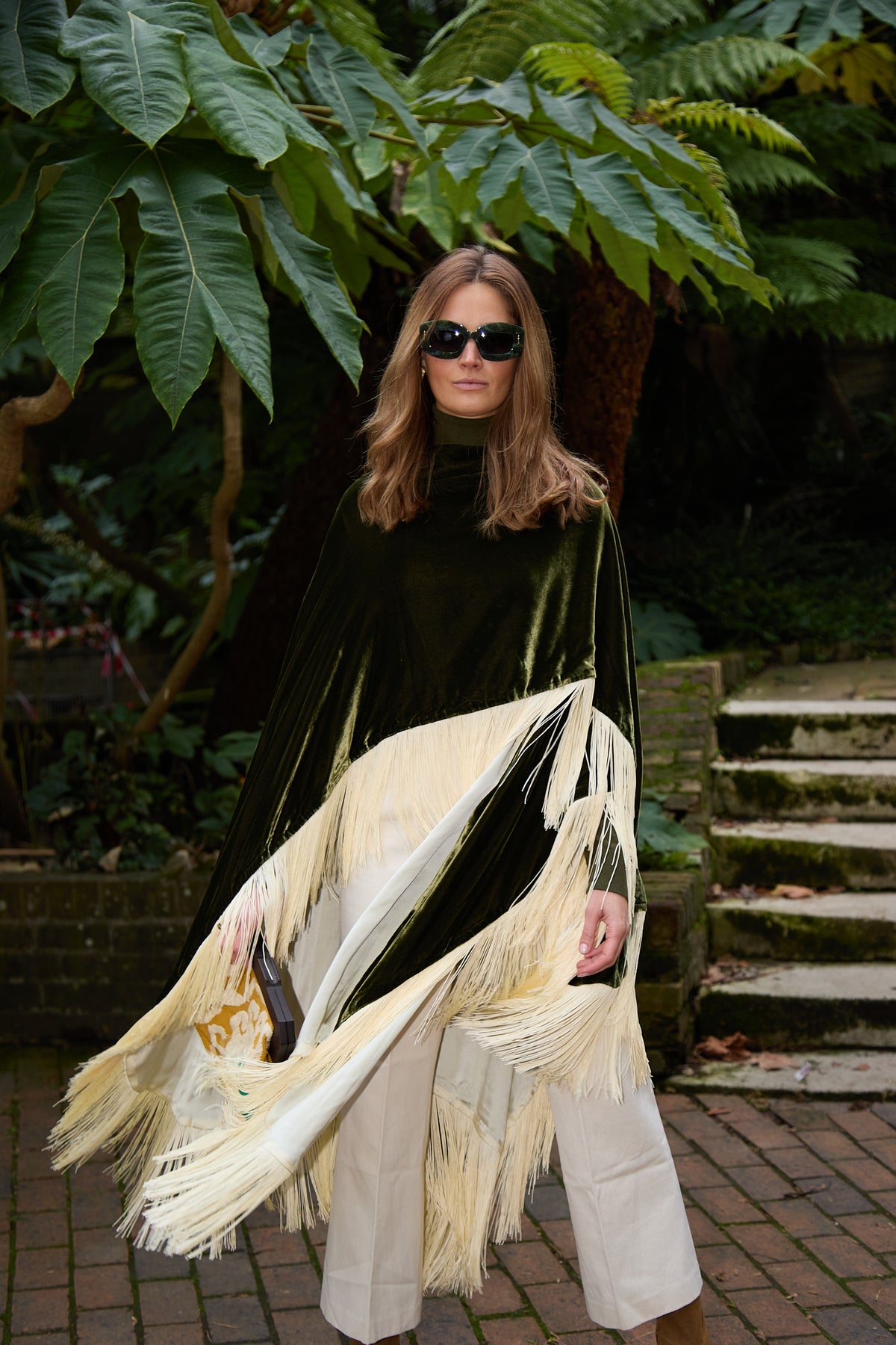 The Evergreen Poncho in olive green silk velvet with cream fringe, front view, garden steps