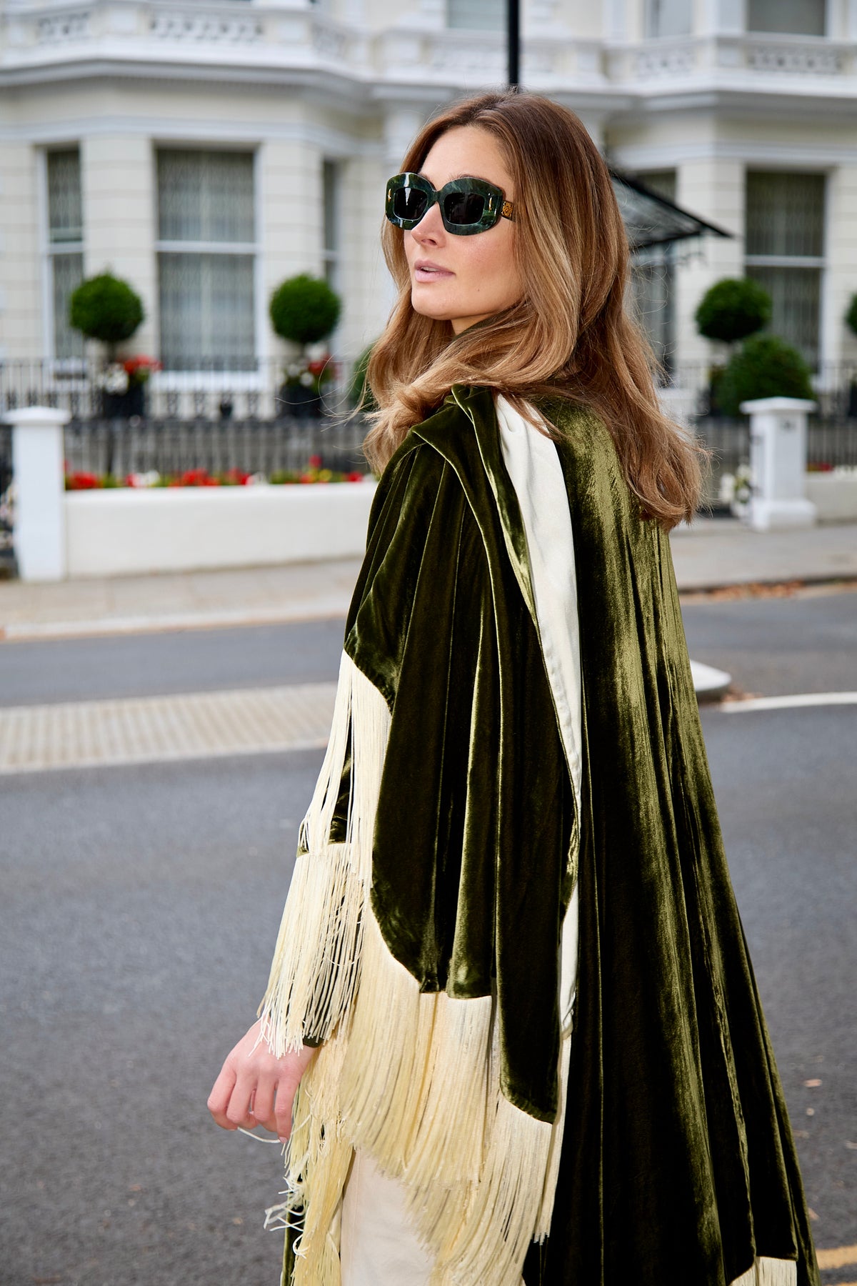 The Evergreen Poncho in olive green silk velvet with cream fringe, side view, London street