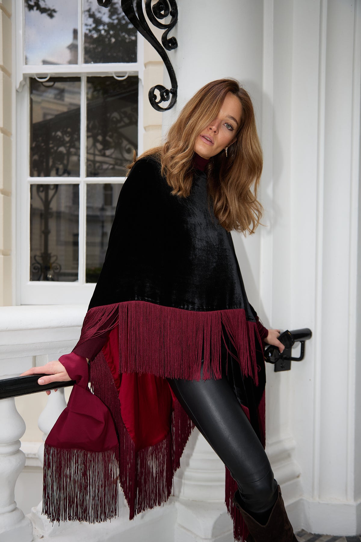 The Black Rose Poncho, short, in black silk velvet with dark red fringe, walking down London townhouse steps