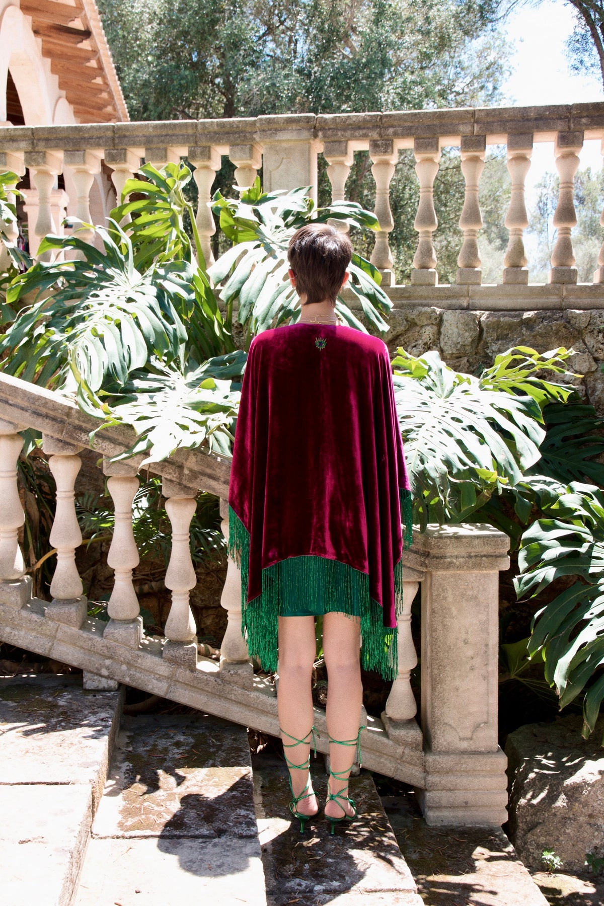 The Dahlia Sparkle Poncho in magenta silk velvet with emerald beaded fringe, back view, garden terrace