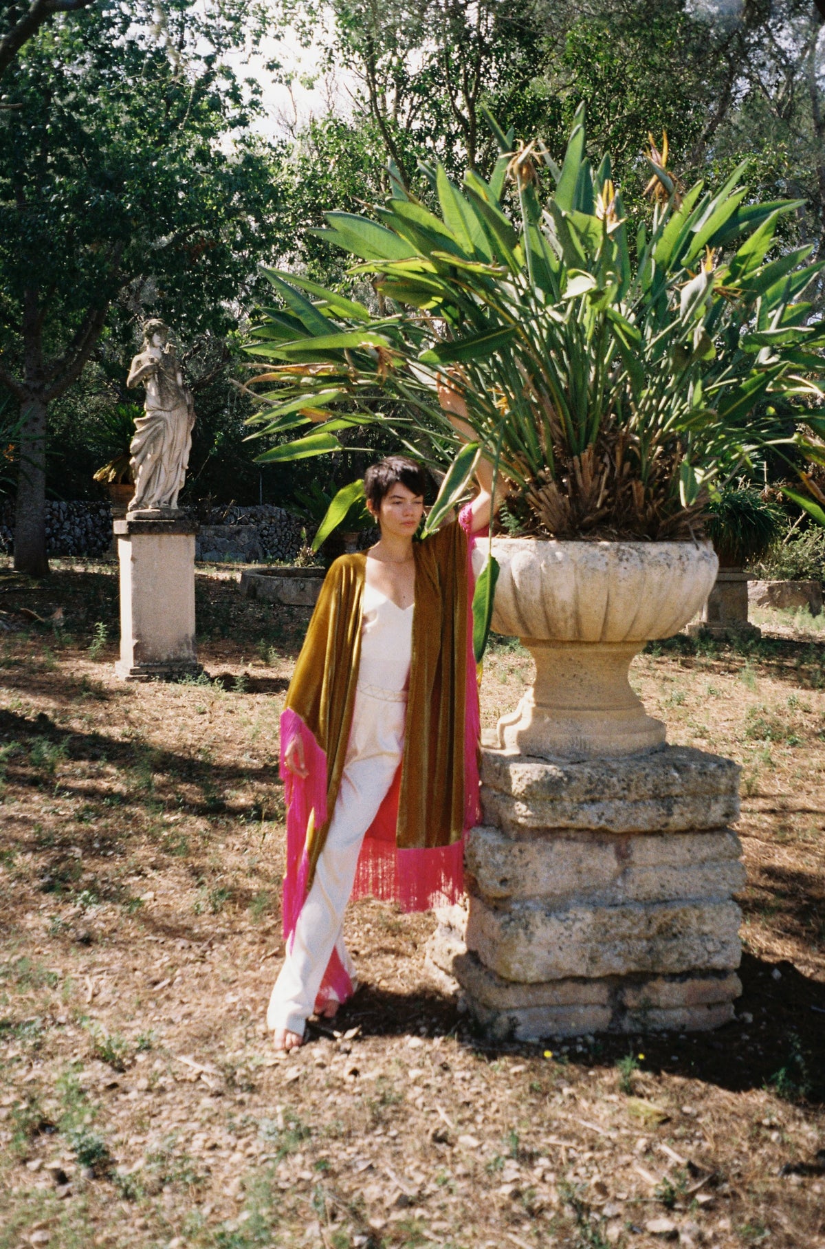 The Limoncello Poncho in golden yellow silk velvet with pink fringe, front view, garden