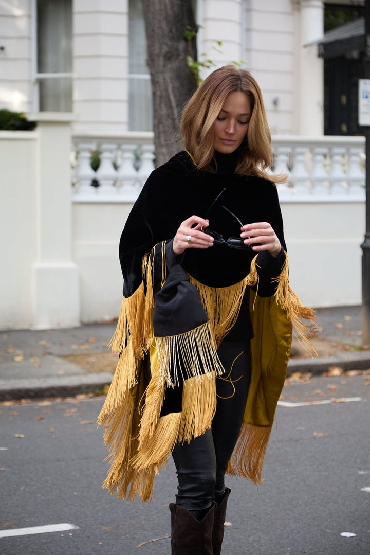 The Black Beaded Gold Knot Bag in black with gold beaded fringe, carried with the Golden Age Poncho, London street