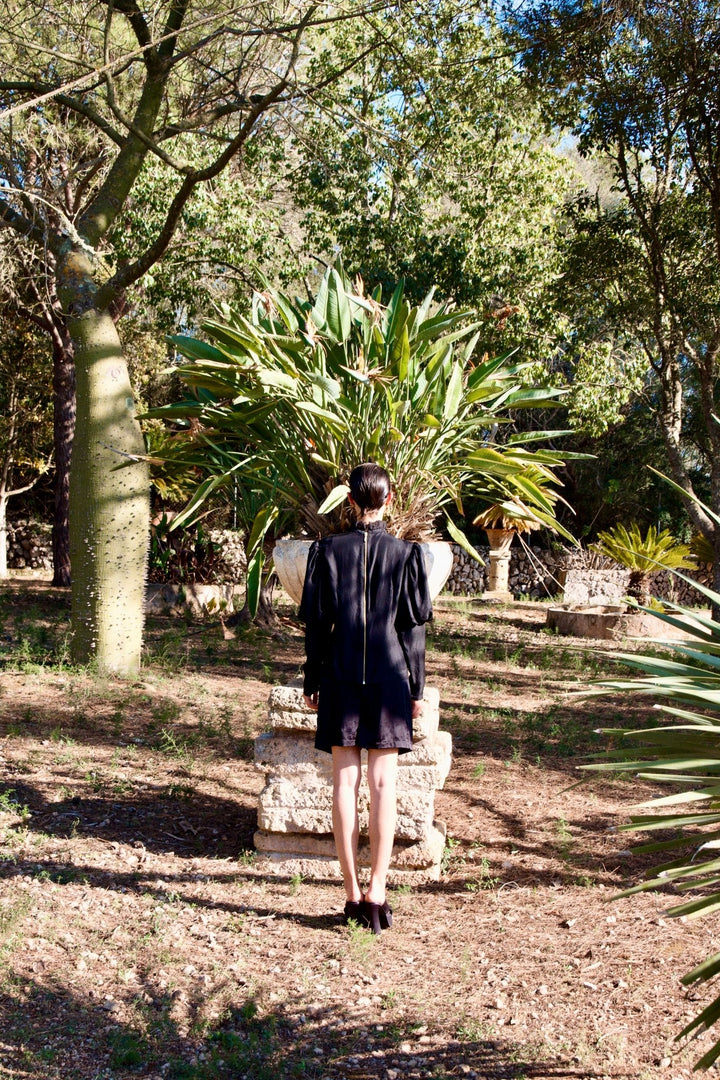 The Black Puff Ruffle Blouse in black silk twill, back view, garden