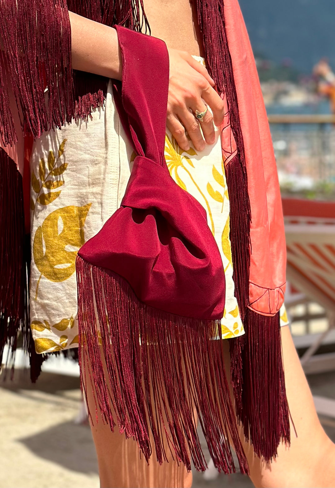 The Burgundy Knot Bag in burgundy silk velvet with long fringe, carried with a coral poncho, seaside