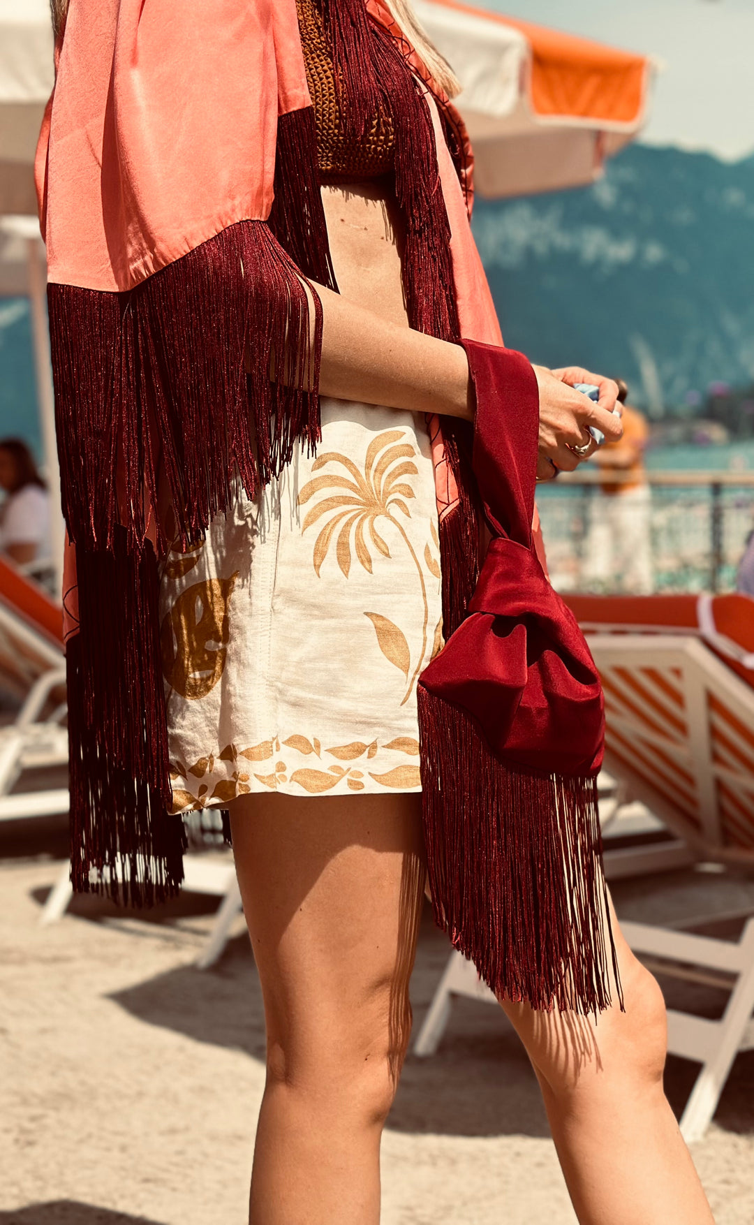 The Burgundy Knot Bag in burgundy silk velvet with long fringe, carried with a coral poncho, seaside
