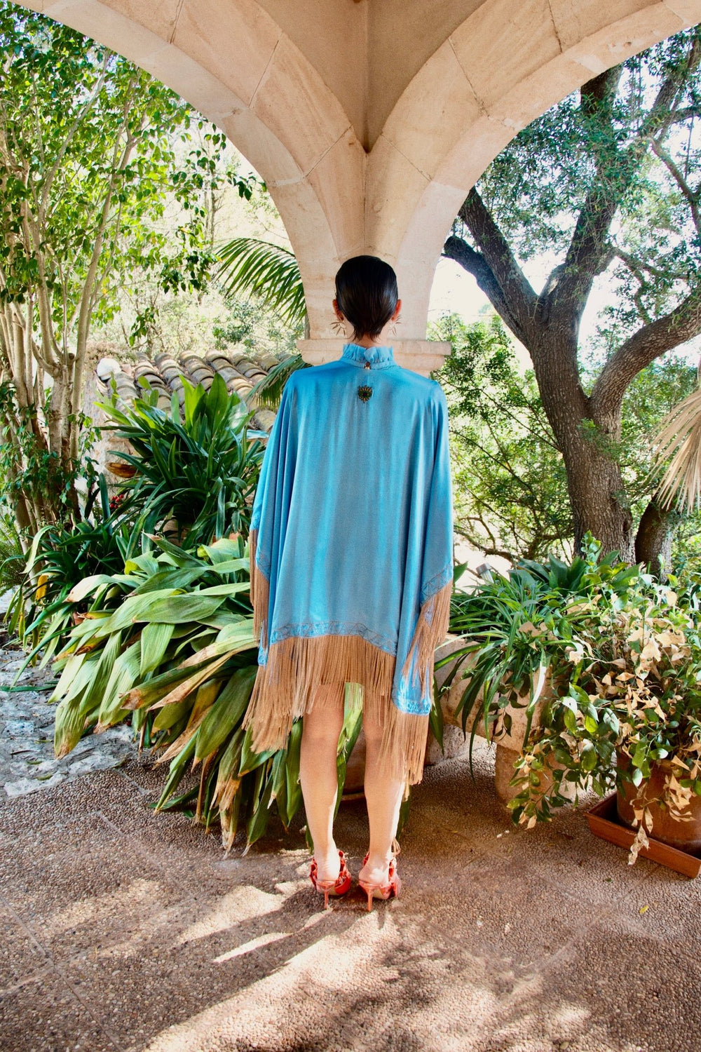 The Cornflower Poncho in powder blue silk with fringed hem, back view, garden archway