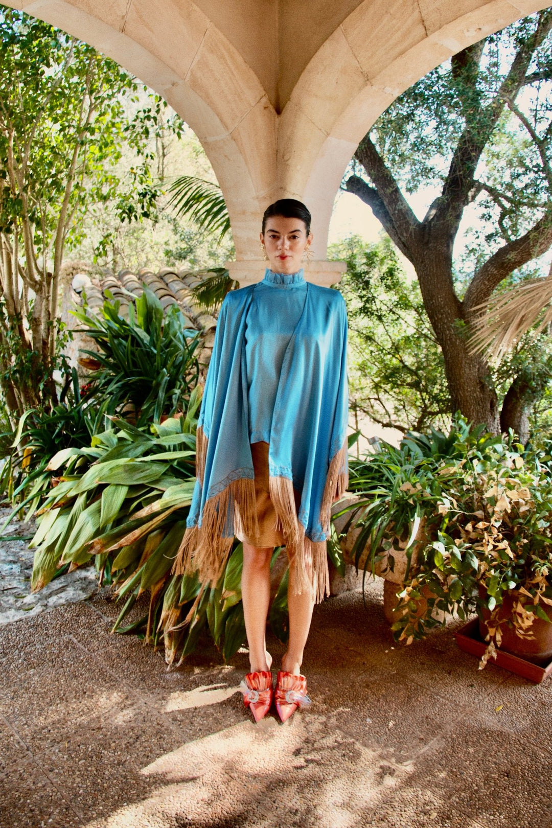 The Cornflower Poncho in powder blue silk with fringed hem, front view, garden archway