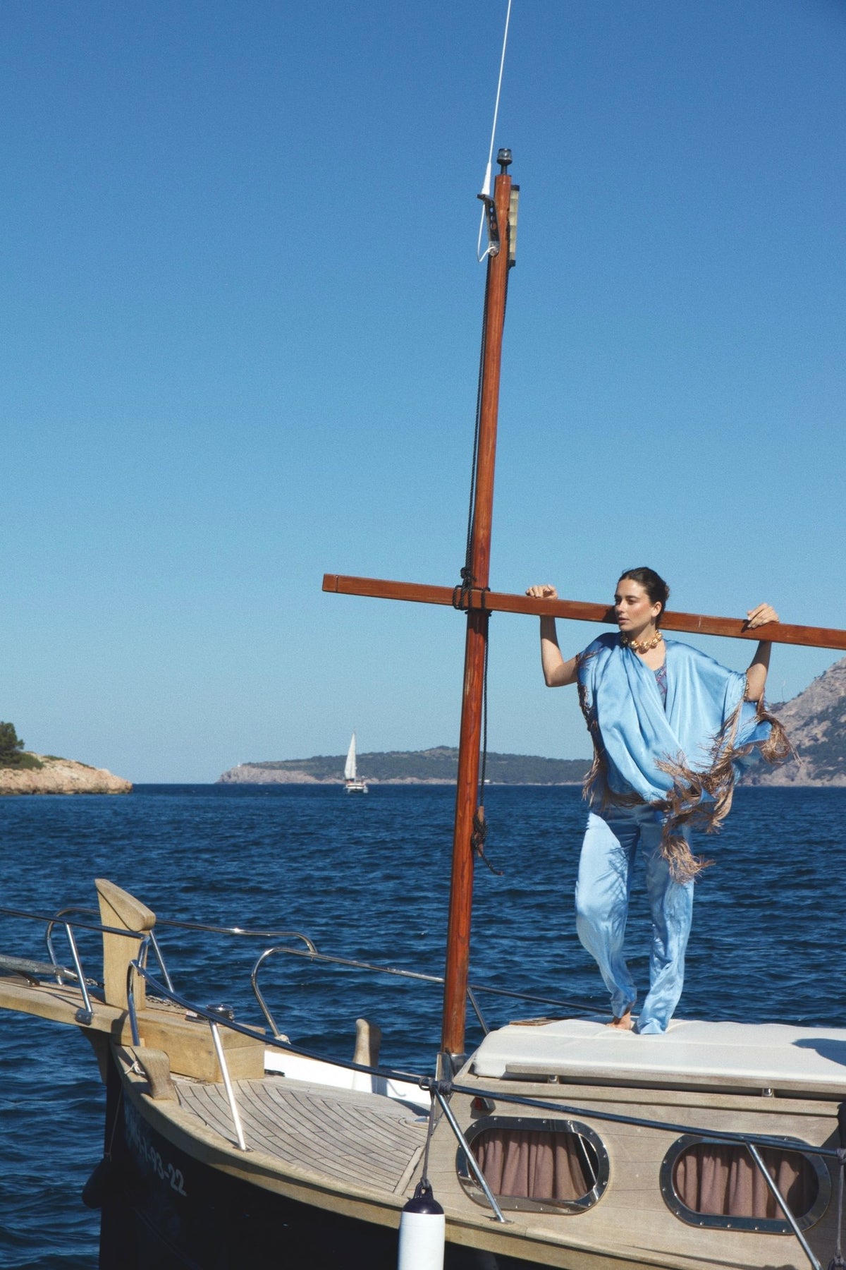 The Cornflower Poncho in powder blue silk with fringed hem, on the deck of a sailing boat