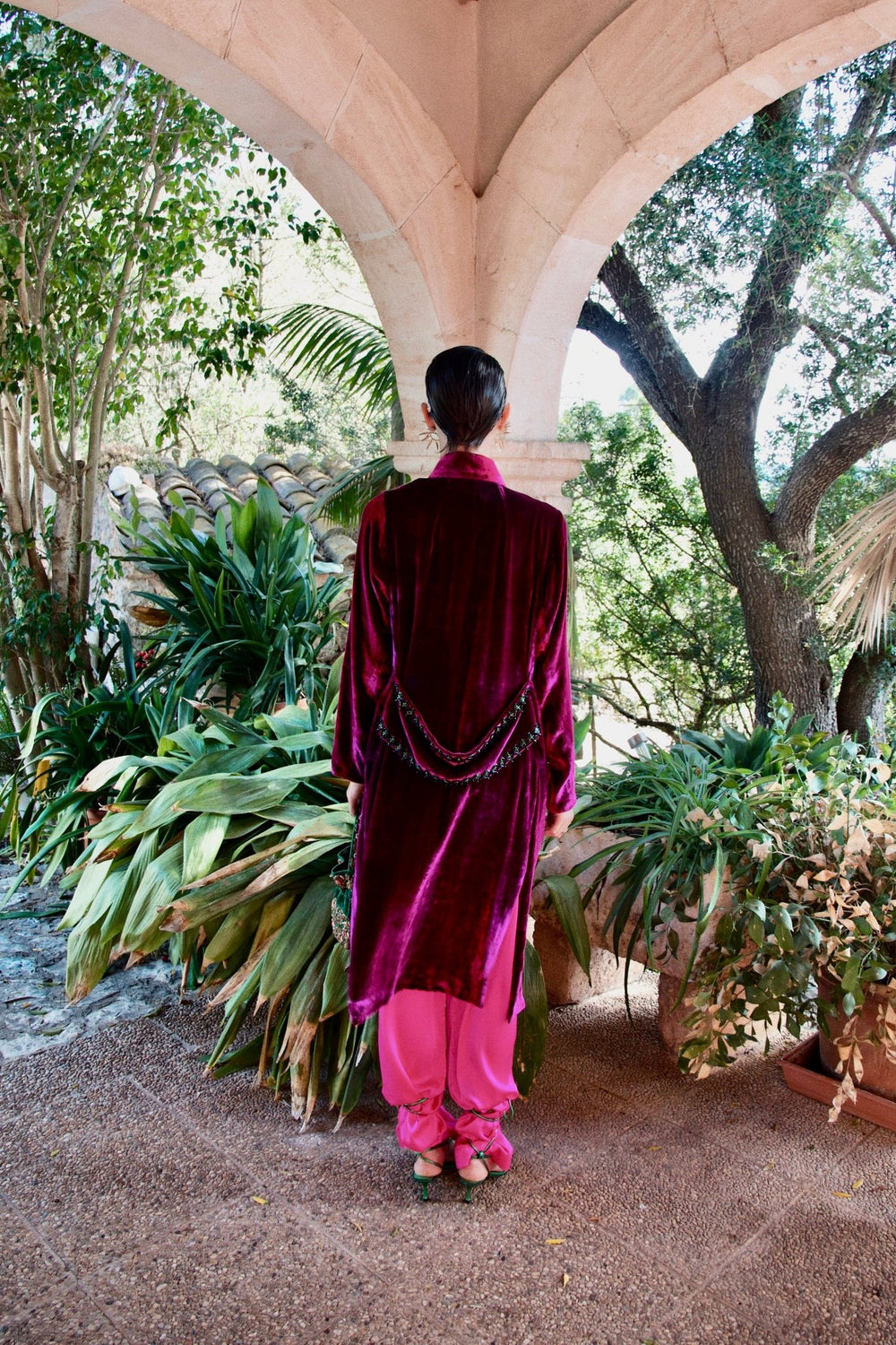 The Dahlia Coat in magenta silk velvet with embroidered trim, back view, garden archway