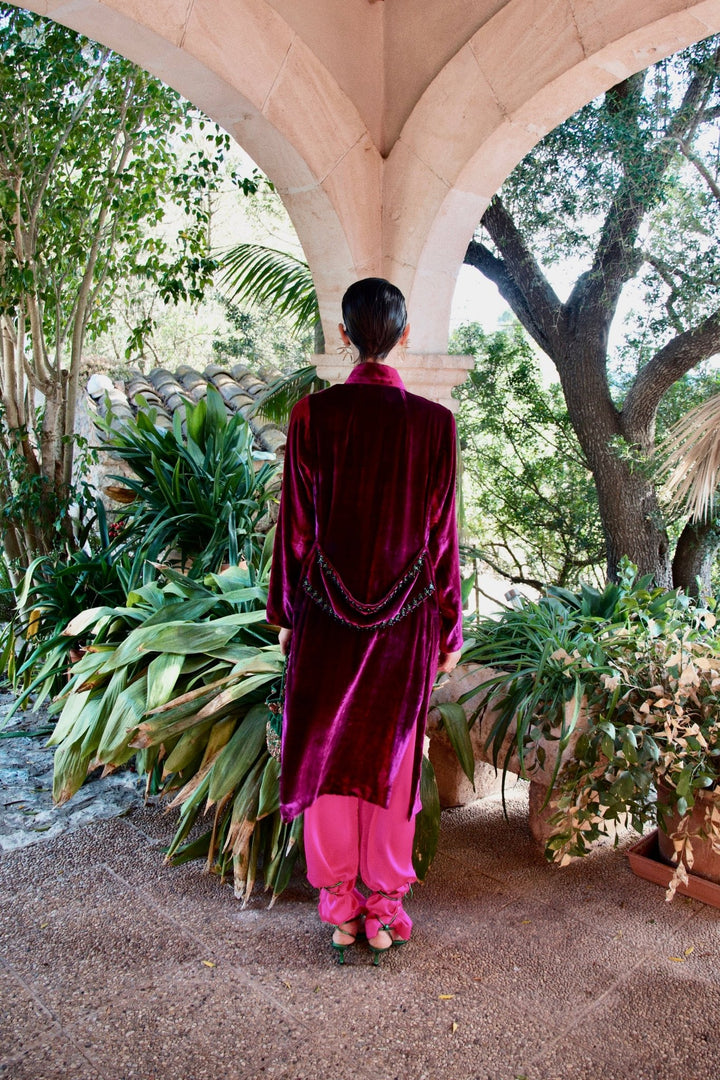The Dahlia Coat in magenta silk velvet with embroidered trim, back view, garden archway