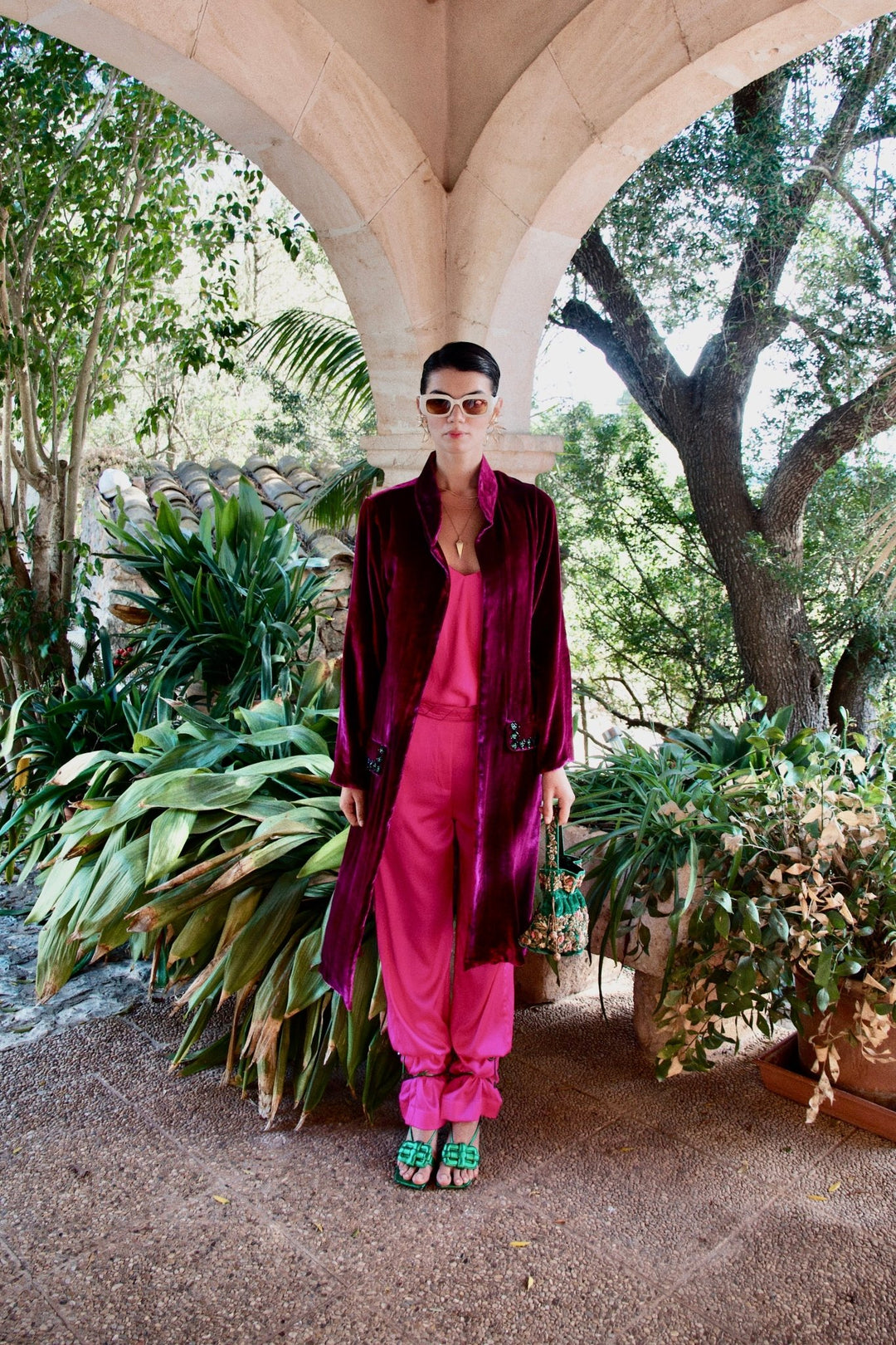 The Dahlia Coat in magenta silk velvet with embroidered trim, front view, garden archway
