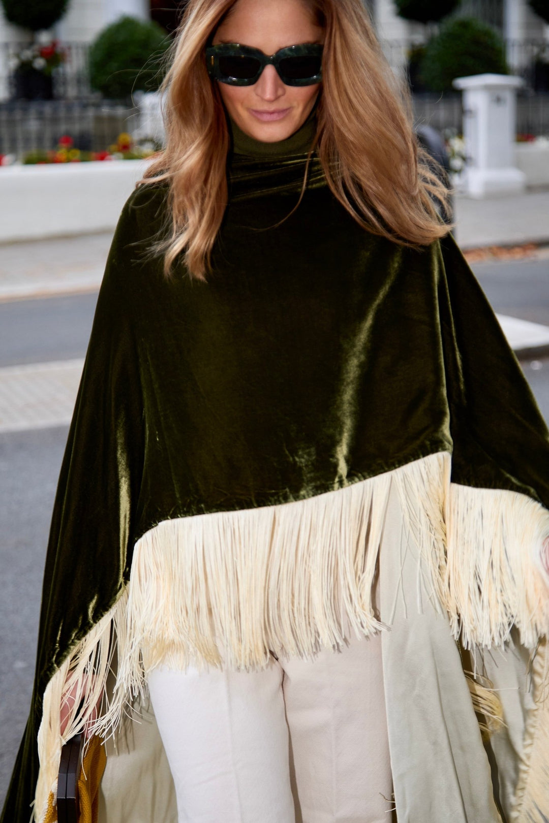 The Evergreen Poncho in olive green silk velvet with cream fringe, close-up, London street
