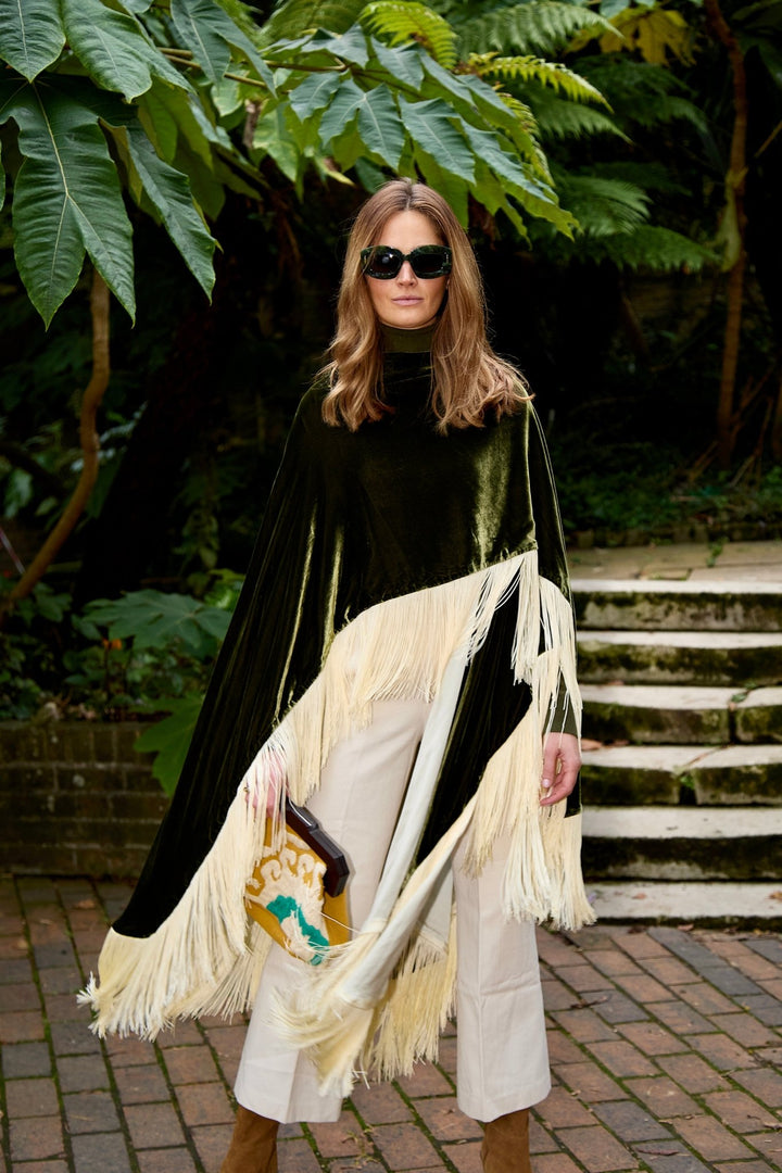 The Evergreen Poncho in olive green silk velvet with cream fringe, front view, garden steps