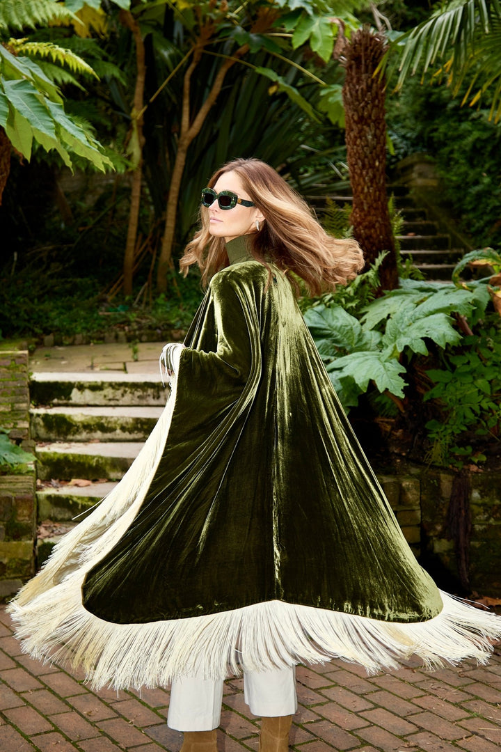 The Evergreen Poncho in olive green silk velvet with cream fringe, in motion, garden steps