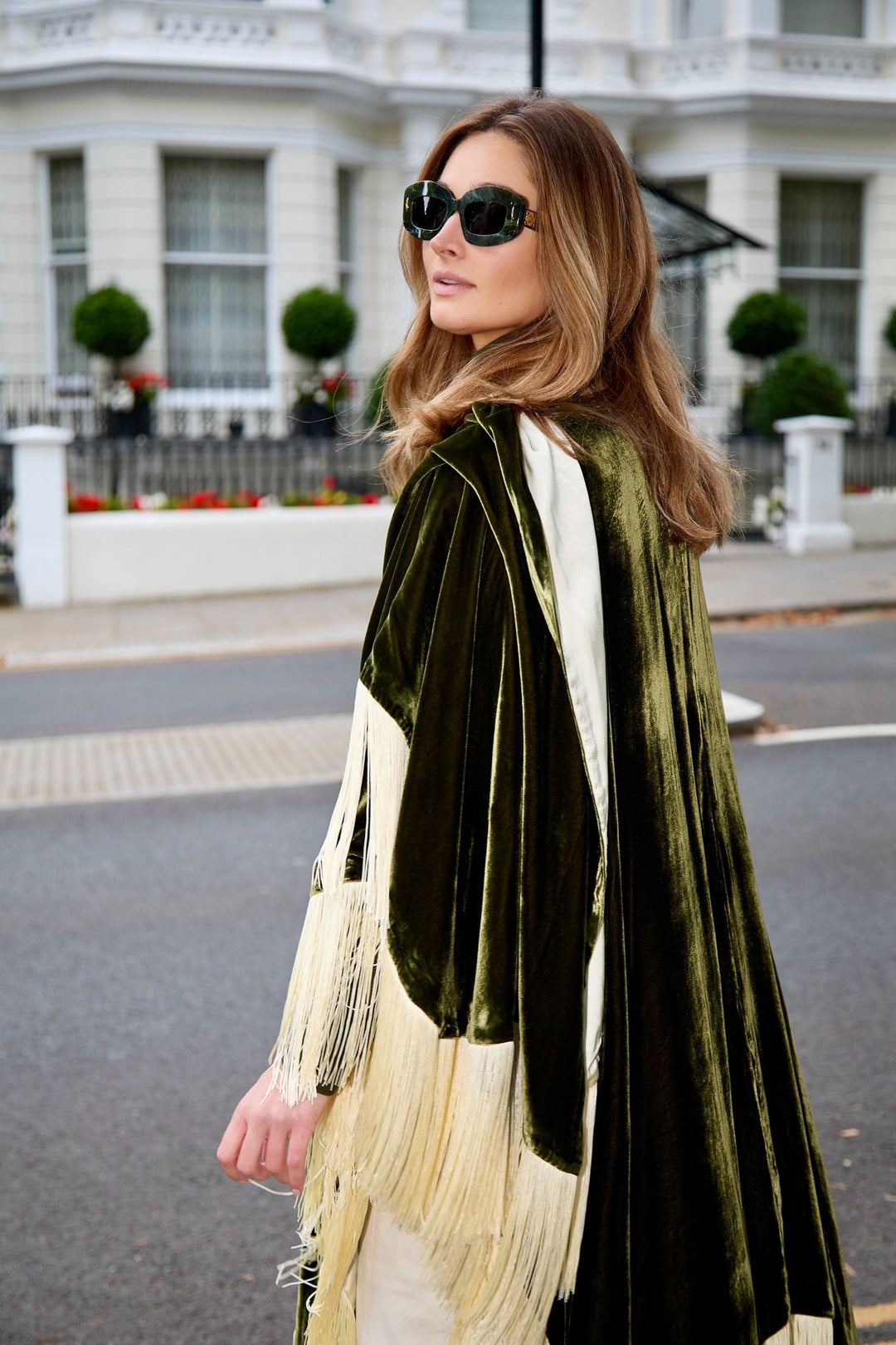 The Evergreen Poncho in olive green silk velvet with cream fringe, side view, London street