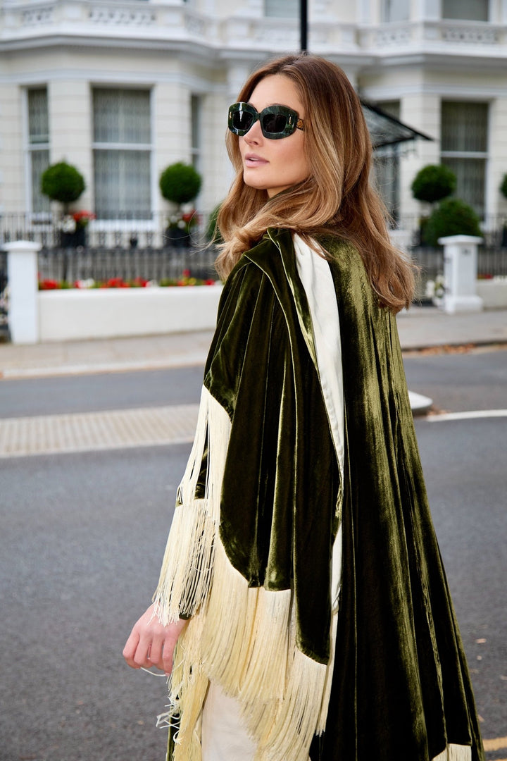 The Evergreen Poncho in olive green silk velvet with cream fringe, side view, London street