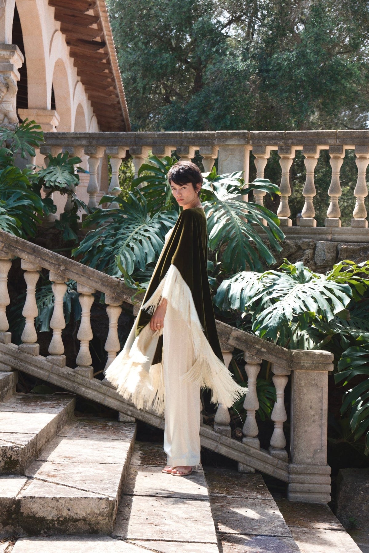The Evergreen Poncho in olive green silk velvet with cream fringe, side view, stone balustrade terrace