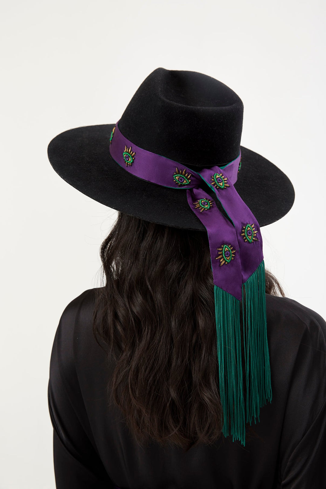 The Eyecatcher Hatband in purple silk with embroidered eyes and green fringe, on a black hat, back view, studio
