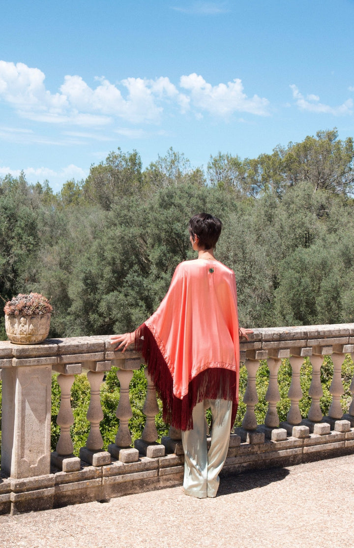 The Fire Bolt Poncho in coral silk with red fringe, back view, stone terrace