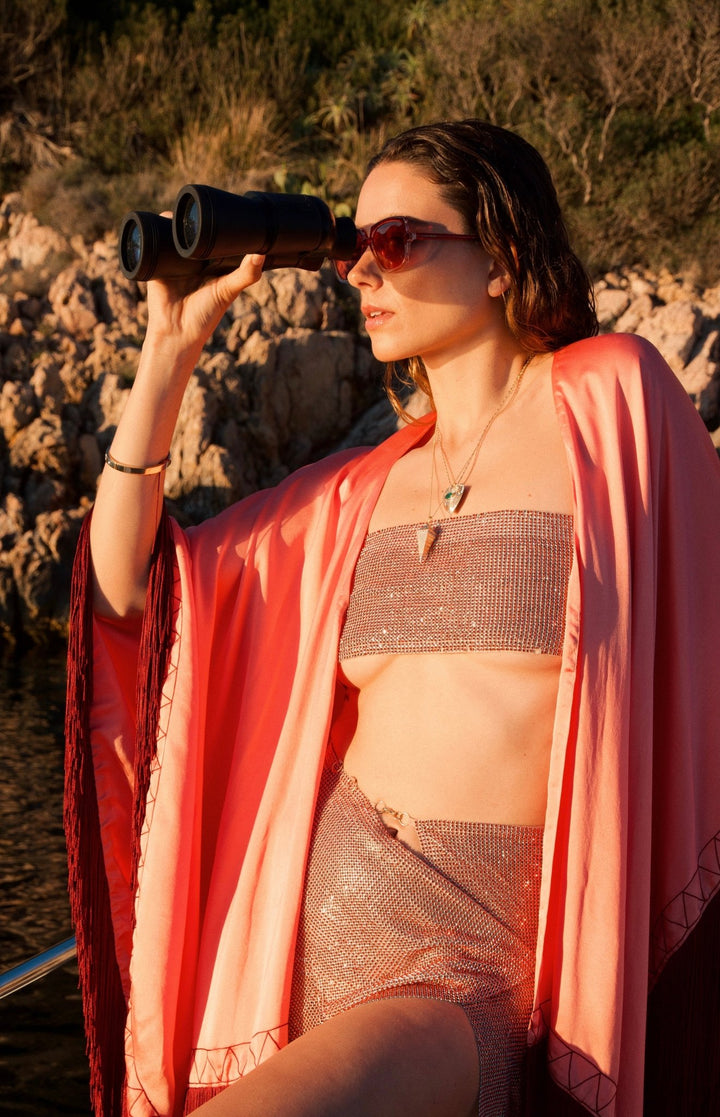 The Fire Bolt Poncho in coral silk with red fringe, close-up, on a sailing boat