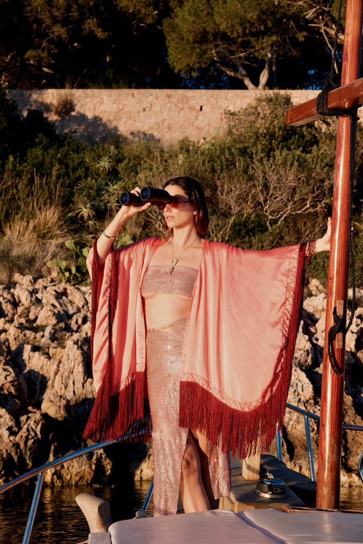 The Fire Bolt Poncho in coral silk with red fringe, front view, on a sailing boat
