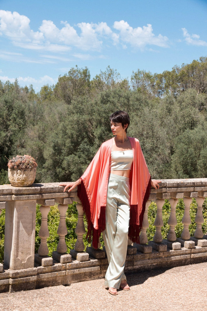 The Fire Bolt Poncho in coral silk with red fringe, front view, stone terrace