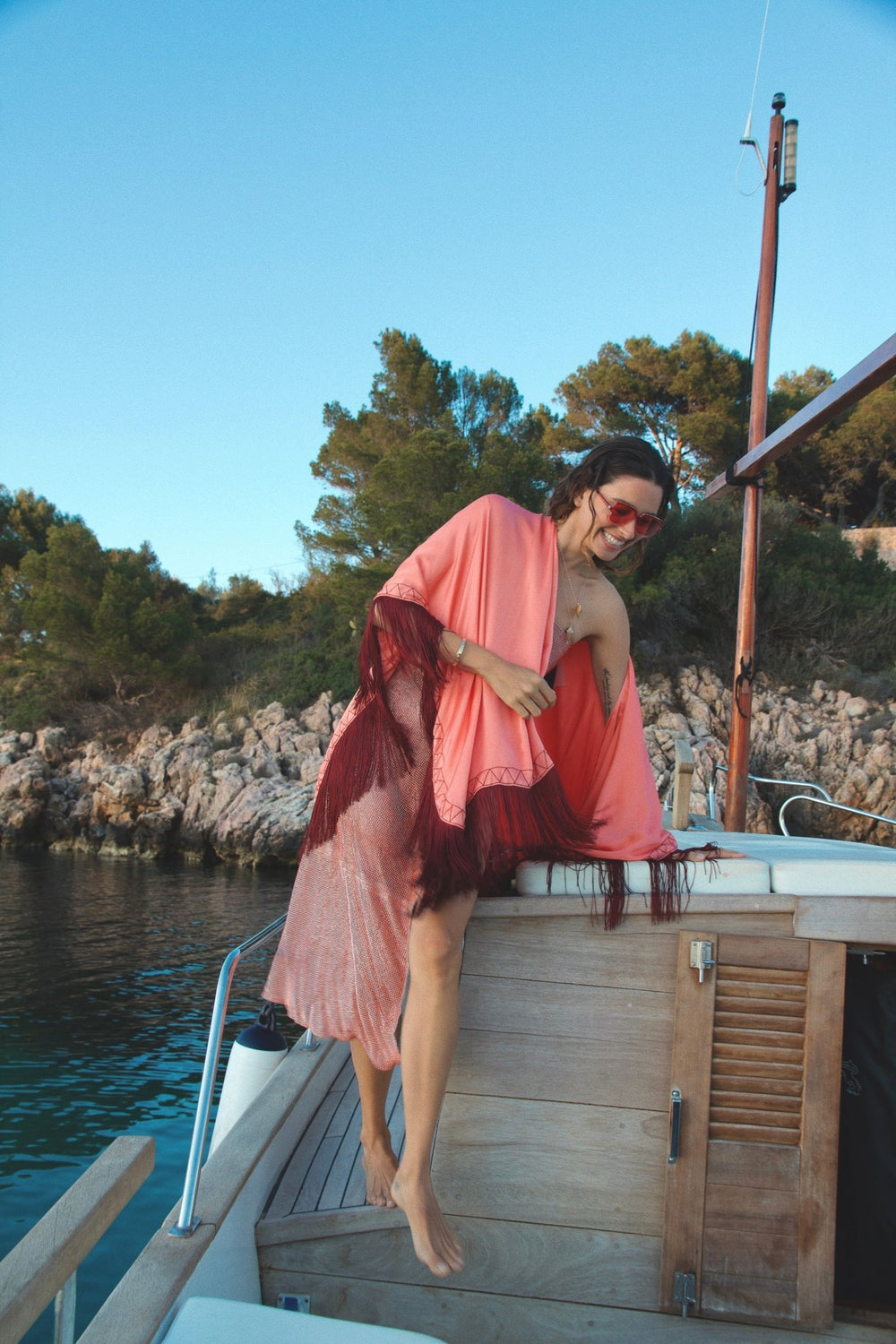 The Fire Bolt Poncho in coral silk with red fringe, on the deck of a sailing boat