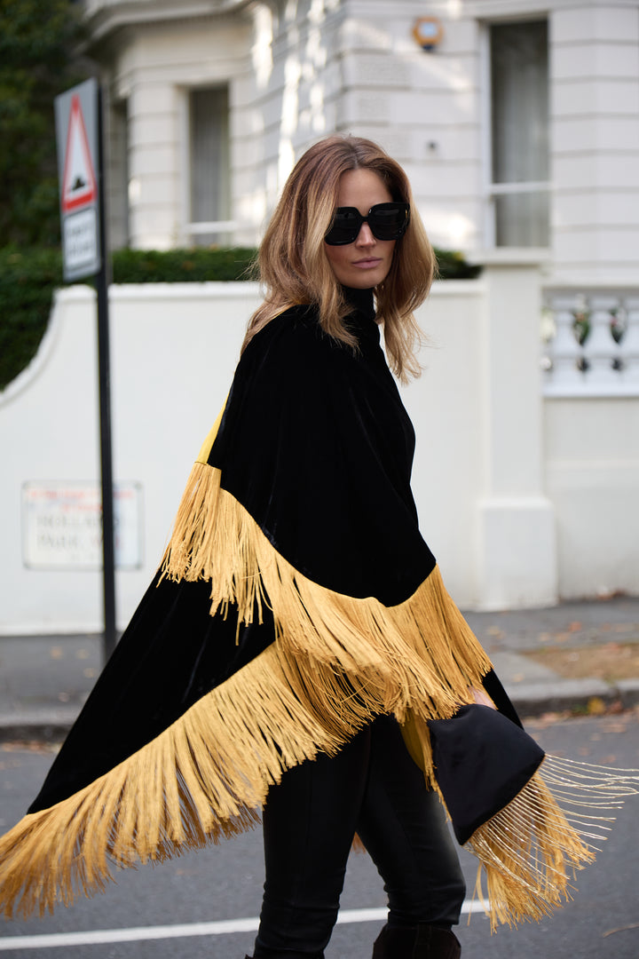 The Golden Age Poncho in black silk velvet with gold fringe, in motion, London street