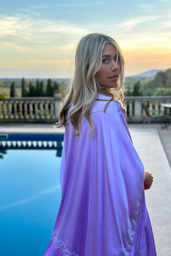 The Lavender Poncho in lavender silk with fringed hem, back view, terrace at sunset