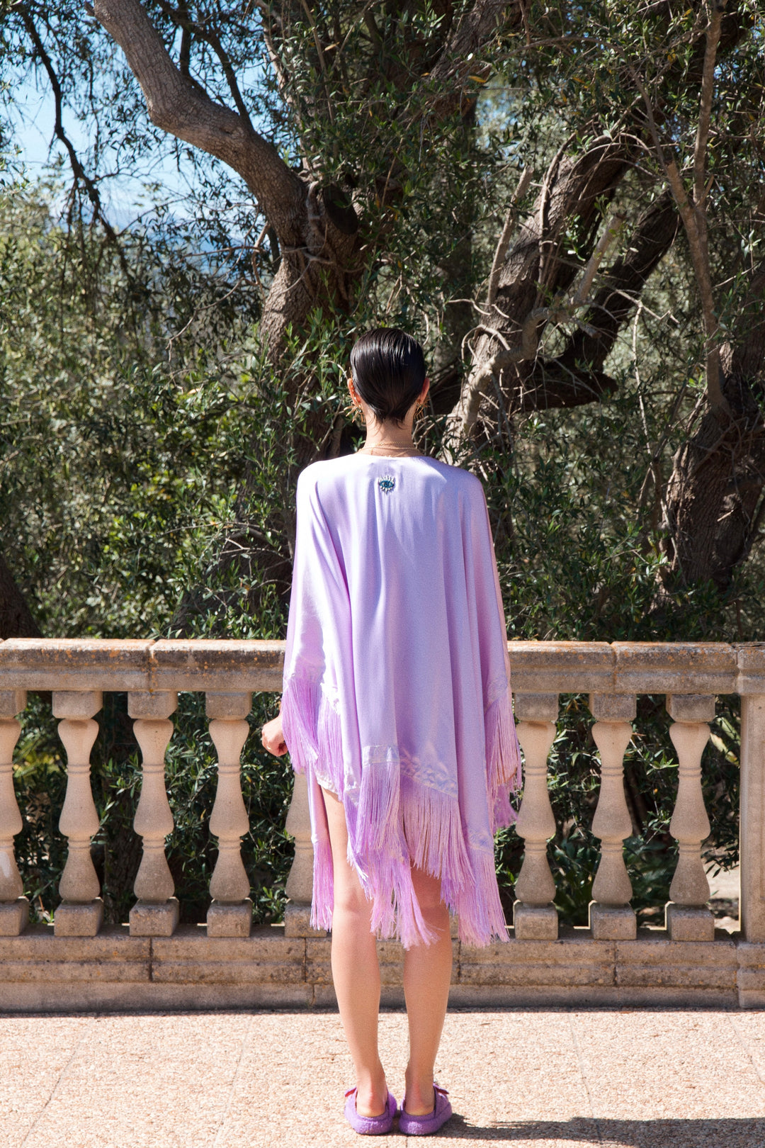 The Lavender Poncho in lavender silk with fringed hem, back view, stone terrace