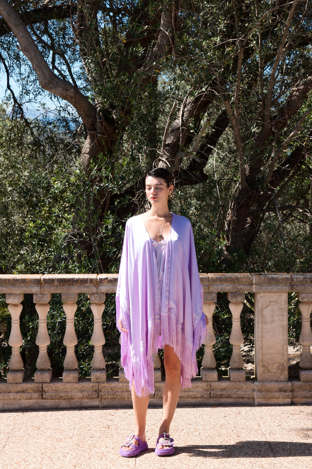 The Lavender Poncho in lavender silk with fringed hem, worn with the Lavender Playsuit, stone terrace