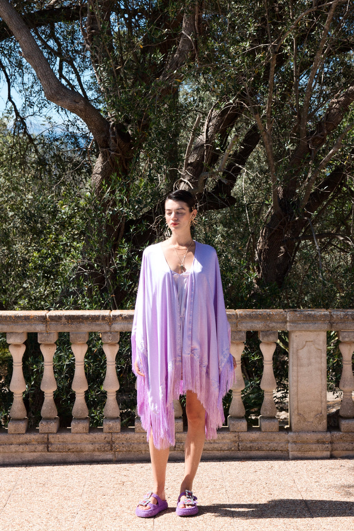 The Lavender Poncho in lavender silk with fringed hem, worn with the Lavender Playsuit, stone terrace