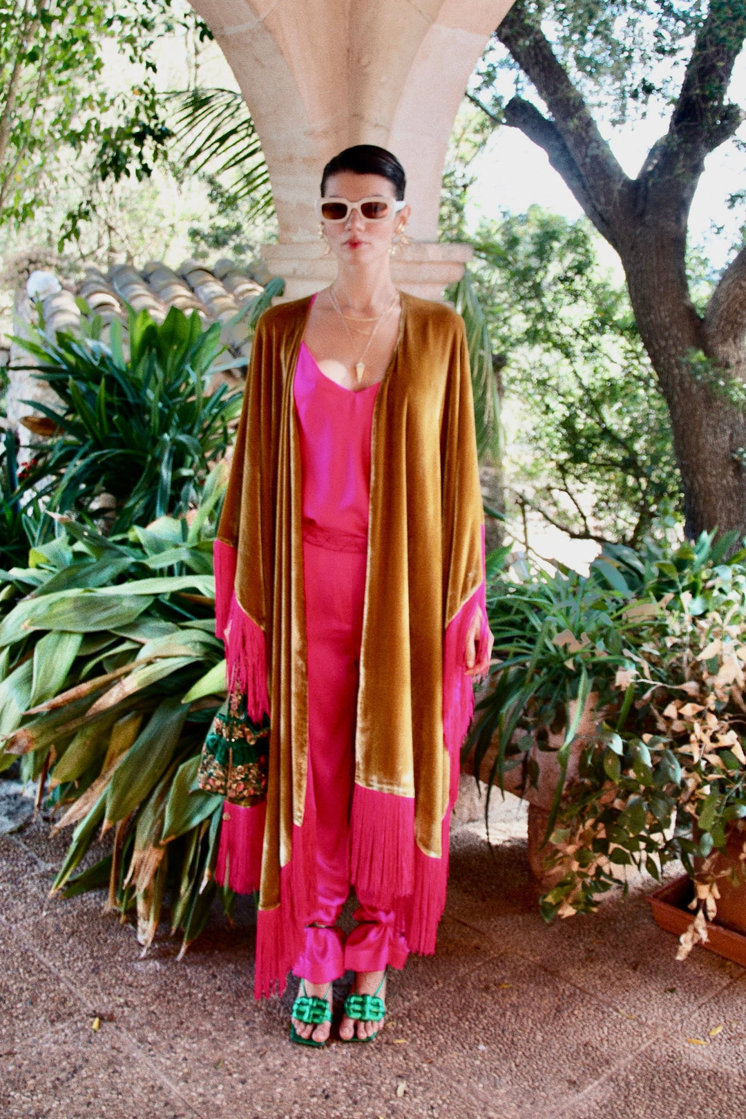 The Limoncello Poncho in golden yellow silk velvet with pink fringe, front view over a pink slip dress, garden archway