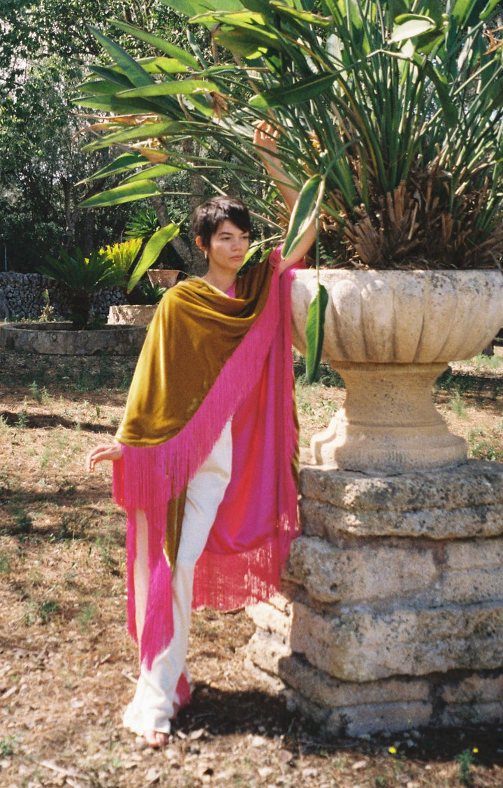 The Limoncello Poncho in golden yellow silk velvet with pink fringe, side view, garden