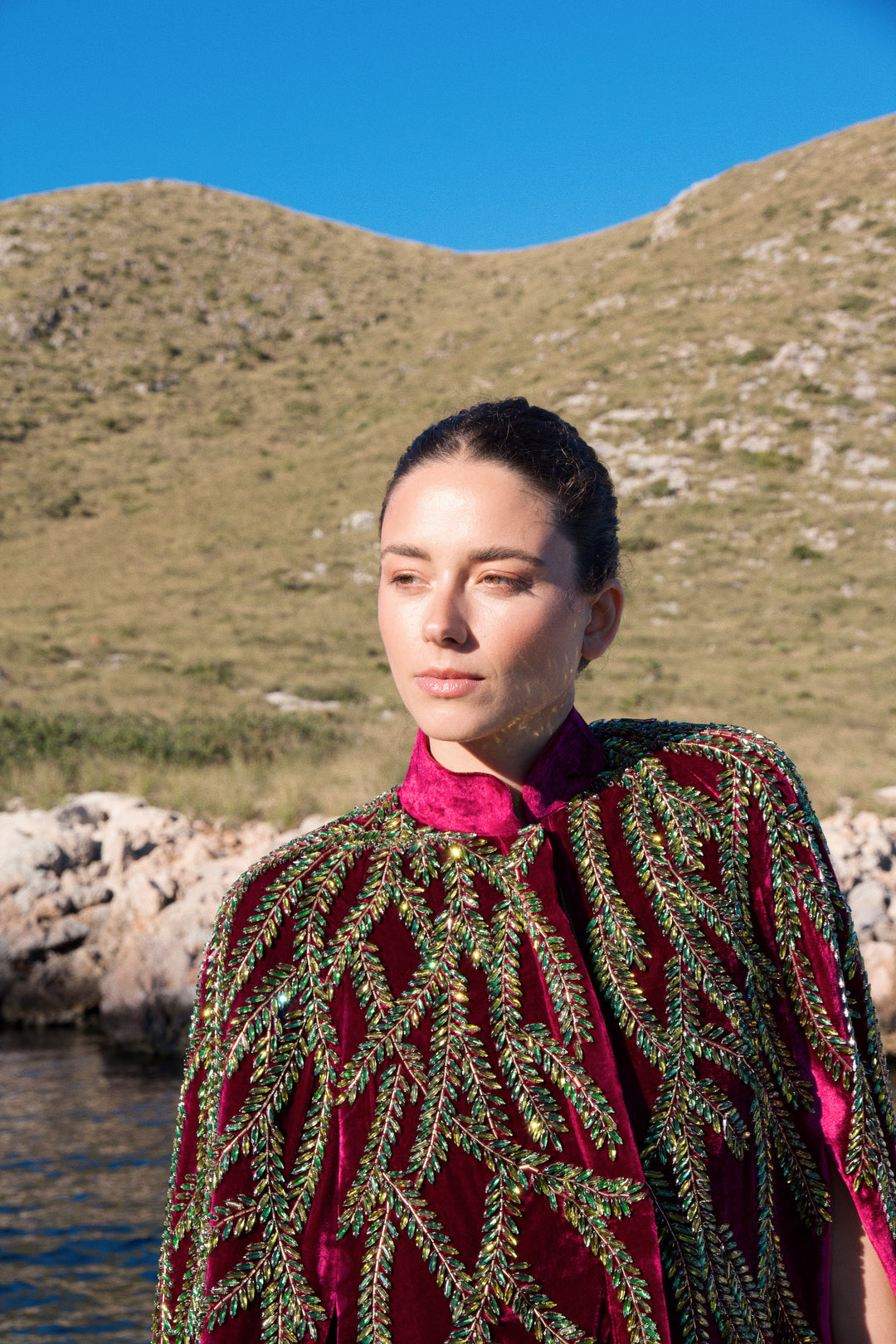 The Royal Garden Cape in magenta silk velvet with gold and green leaf embroidery, close-up, coastal hillside