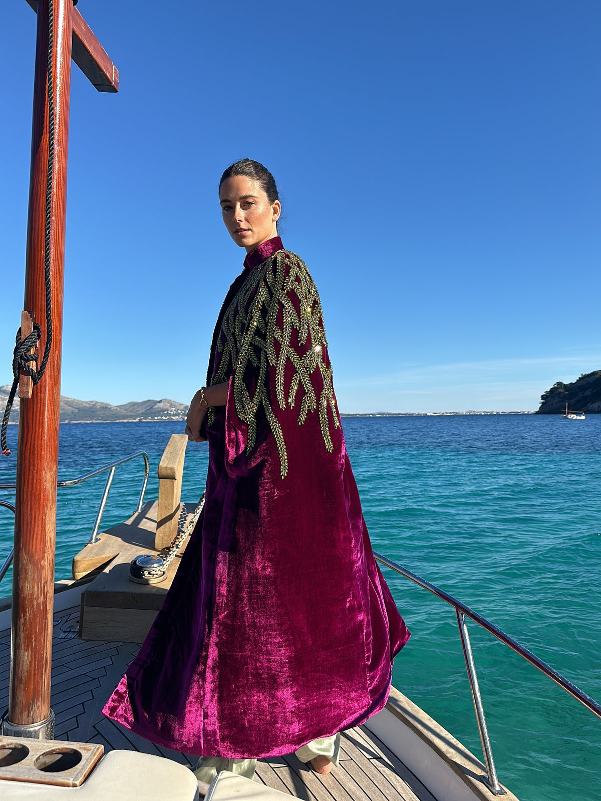The Royal Garden Cape in magenta silk velvet with gold and green leaf embroidery, side view, on a boat at sea
