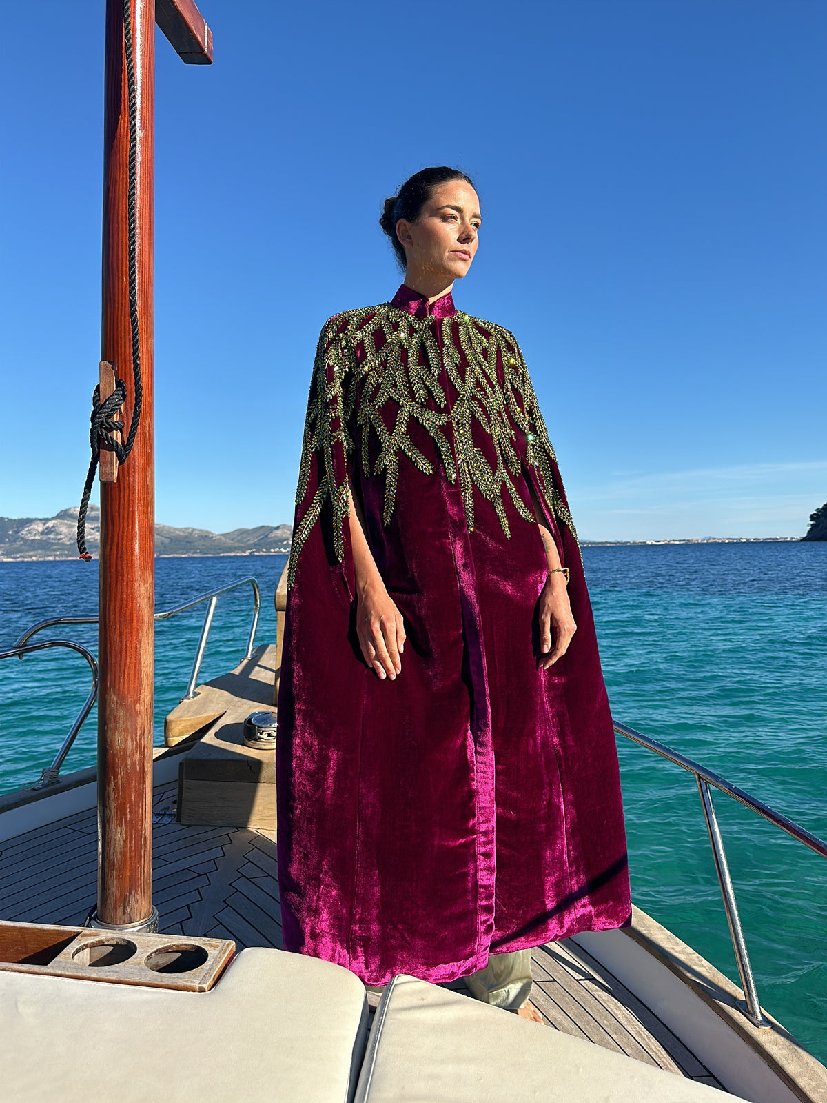 The Royal Garden Cape in magenta silk velvet with gold and green leaf embroidery, front view, on a boat at sea