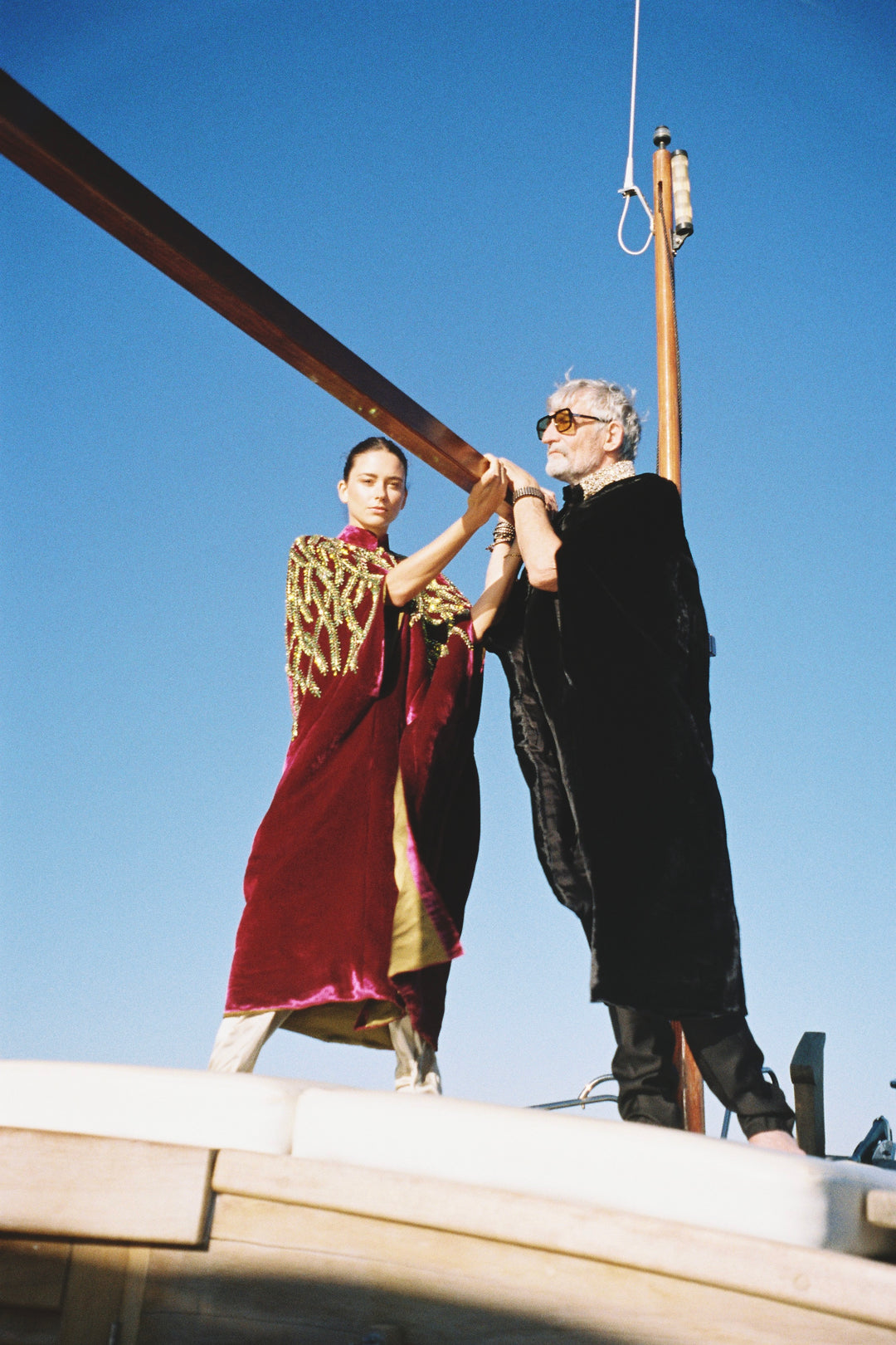 The Royal Garden Cape in magenta silk velvet with gold and green leaf embroidery, on the deck of a sailing boat
