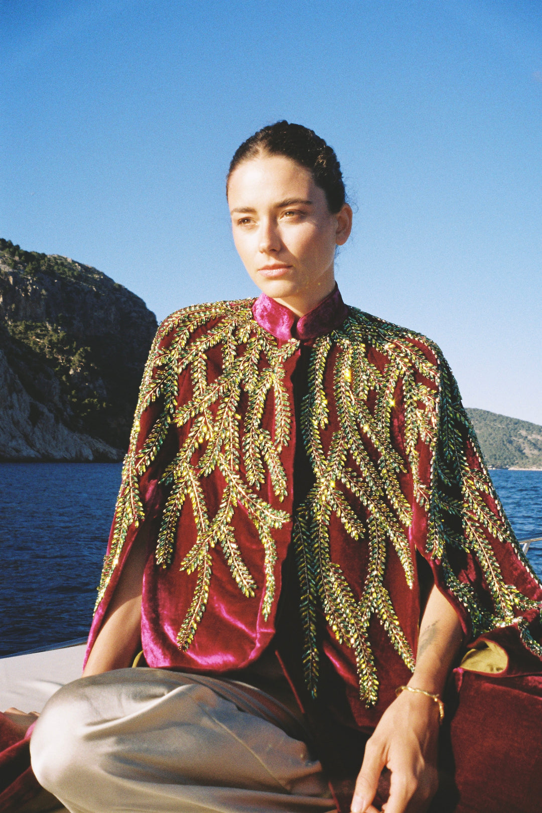 The Royal Garden Cape in magenta silk velvet with gold and green leaf embroidery, seated on a boat at sea