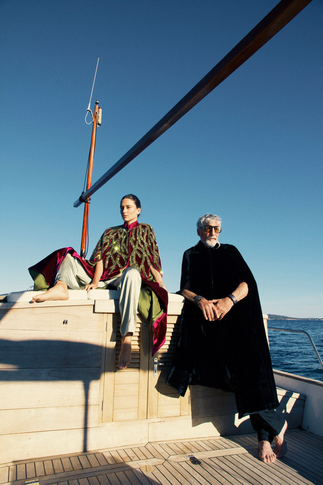 The Royal Garden Cape in magenta silk velvet with gold and green leaf embroidery, seated on a sailing boat