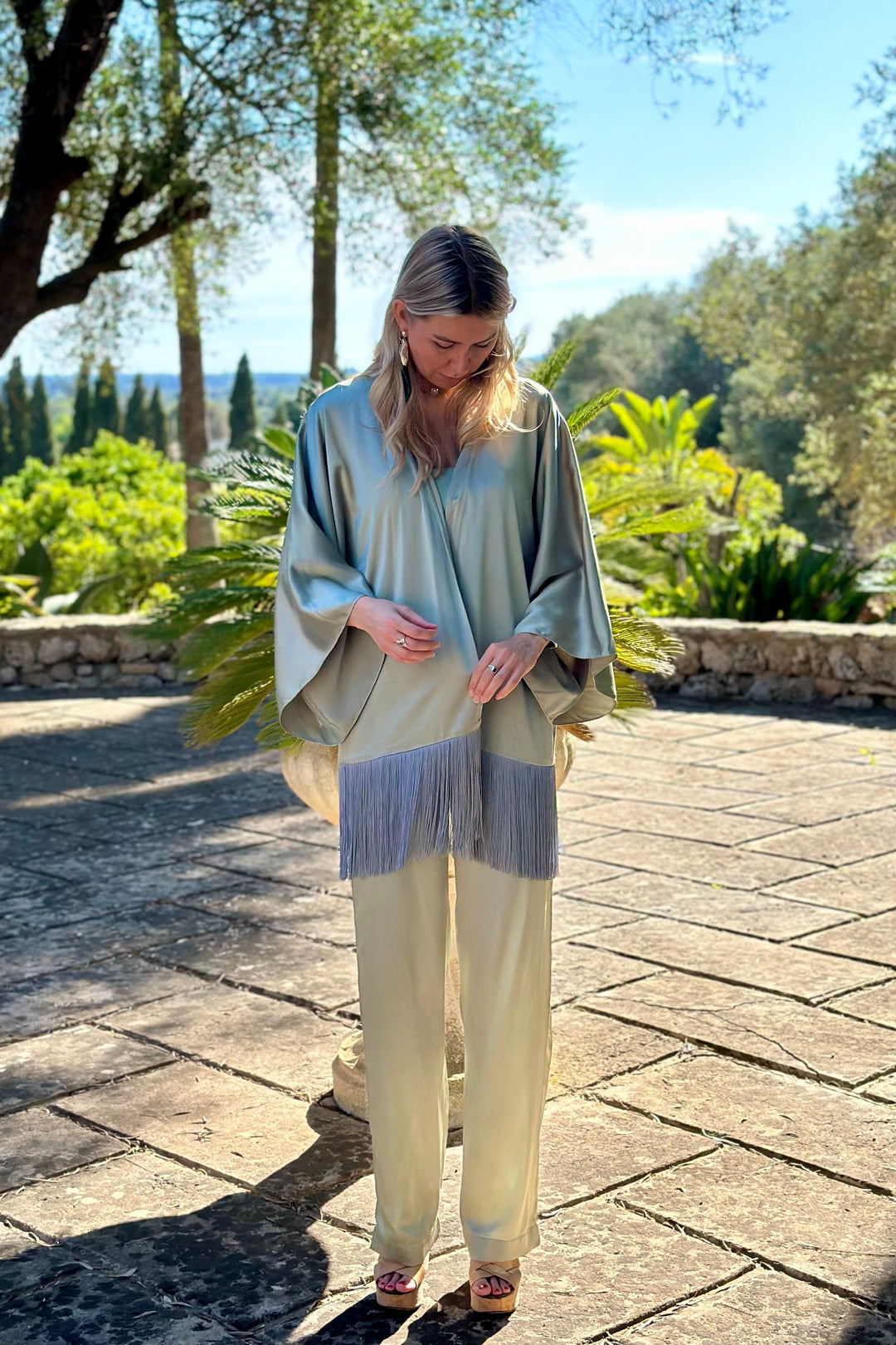 The Sage Leaf Kimono in sage green silk with fringe, front view with matching trousers, terrace