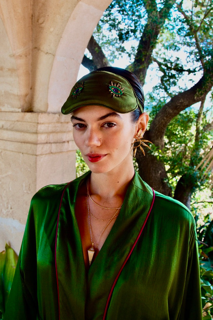 The Silk Sleeping Mask "Eyes Wide Open" in olive green silk with embroidered eyes, worn as a headband, garden archway