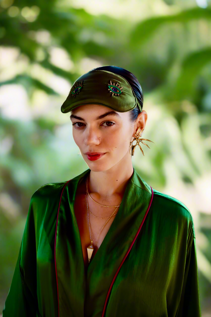 The Silk Sleeping Mask "Eyes Wide Open" in olive green silk with embroidered eyes, worn as a headband, garden