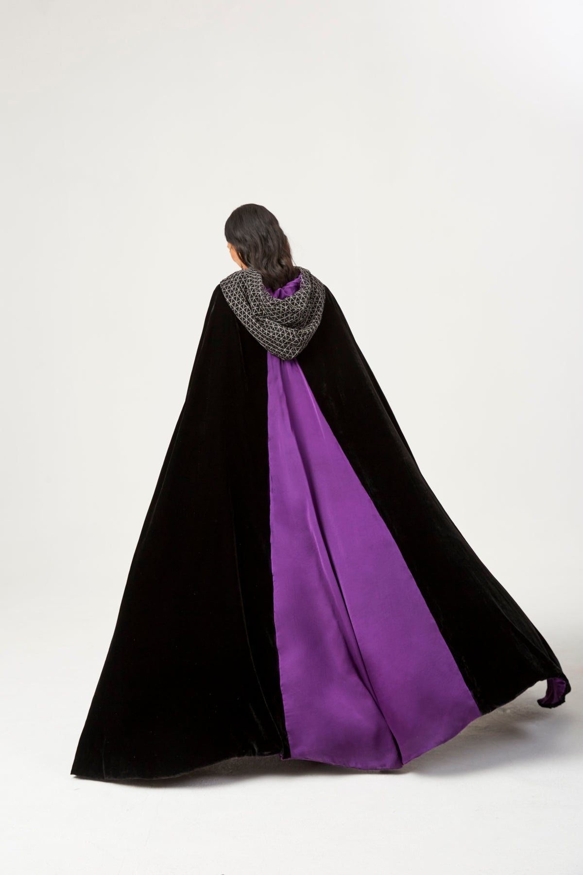 The Mystery Cape in black silk velvet with purple lining and embroidered hood, back view in motion, studio